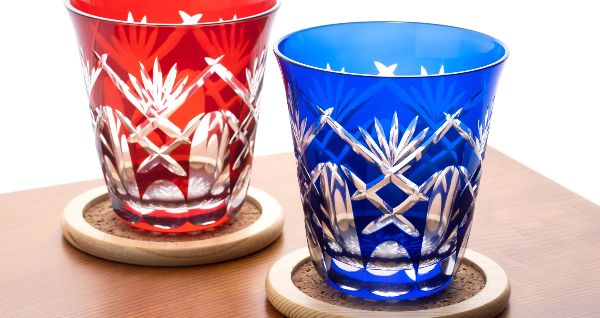 Two cut-glass tumblers, one red and one blue, with intricate geometric patterns, resting on wooden coasters.