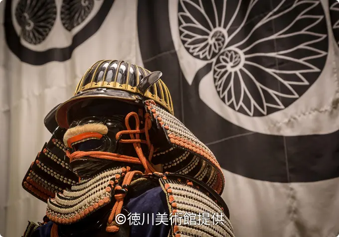 Close-up of a samurai helmet (kabuto) and face guard (menpo) armor, displayed in front of a banner with the Tokugawa family crest.