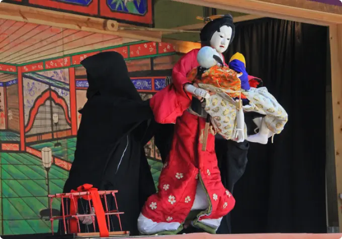 A traditional Japanese puppet show featuring a female puppet in a red kimono holding a smaller puppet, manipulated by two puppeteers in black robes (kuroko) on a stage.