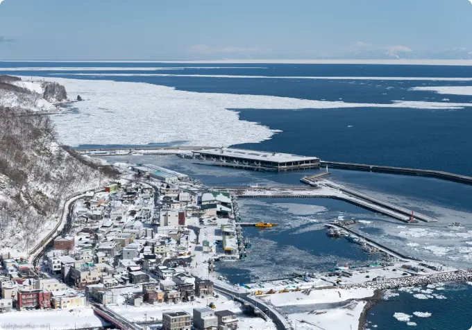 Aerial winter view of a snowy Nemuro, Hokkaido, harbor and town, with large fields of drift ice (Ryūhyō) floating on the Okhotsk Sea in the background.
