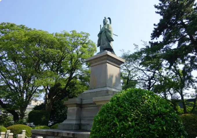 Bronze statue of Ii Naosuke, a feudal lord, standing on a stone pedestal in a lush green park on a sunny day.