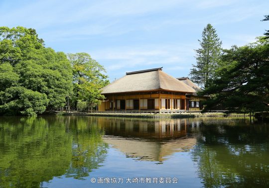 Traditional Japanese building with a thatched roof reflected on a pond, surrounded by lush green trees.