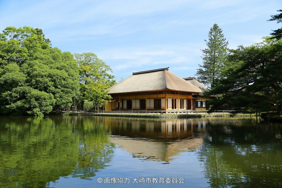 Traditional Japanese building with a thatched roof reflected on a pond, surrounded by lush green trees.
