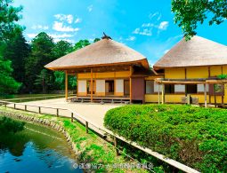 Two traditional Japanese thatched-roof buildings by a pond in a bright green garden under a blue sky.