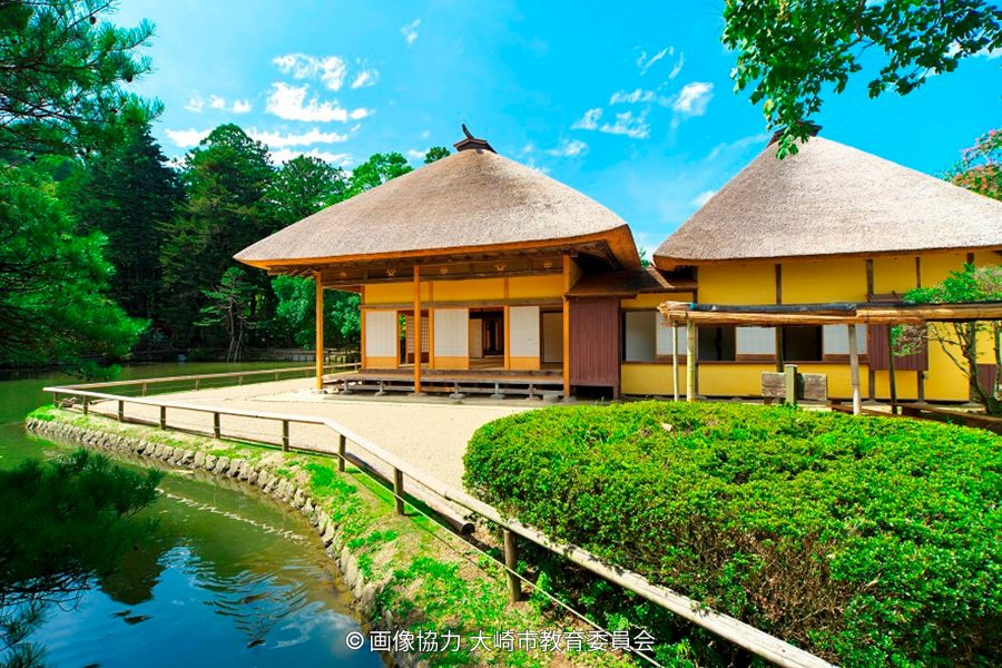 Two traditional Japanese thatched-roof buildings by a pond in a bright green garden under a blue sky.