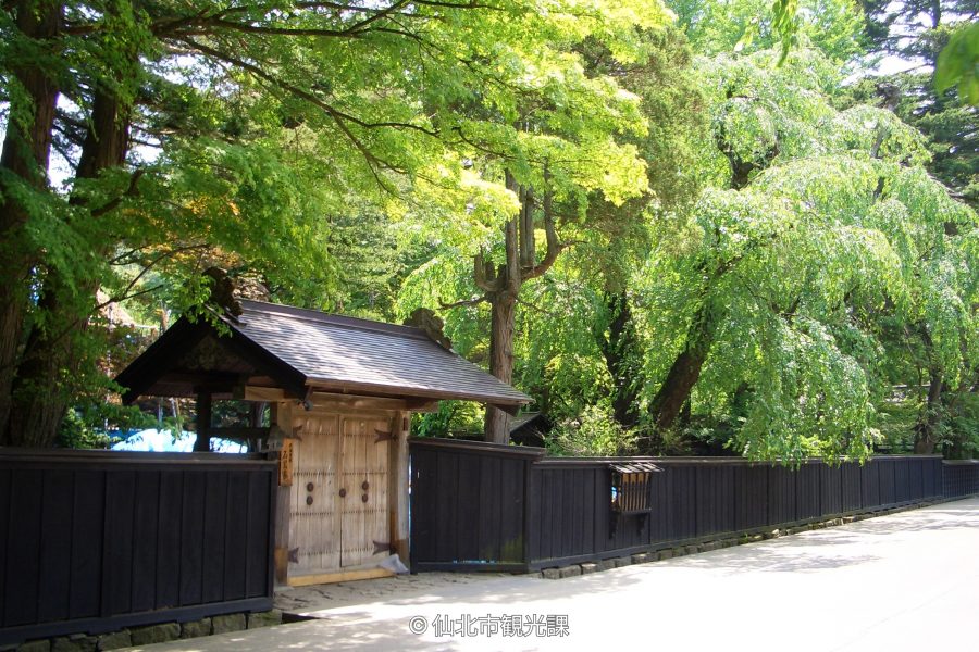 Samurai residence gate and black wooden fence on Kakunodate Street, surrounded by fresh green foliage.