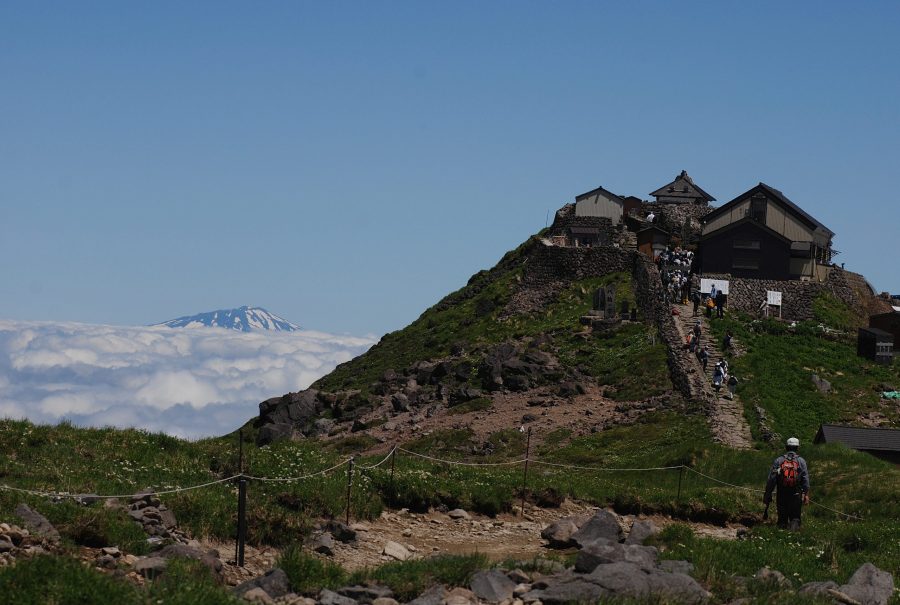 A shrine complex on a mountain summit above the clouds, with a winding stone path leading up to the buildings. (Likely Mt. Gassan Shrine or the Sanjin Gosaiden approach on Mt. Haguro).