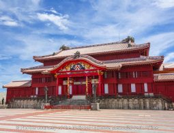 The majestic Seiden (Main Hall) of Shuri Castle, a vibrant red, Okinawan-style wooden building with a tiled plaza in front, set against a bright blue sky.