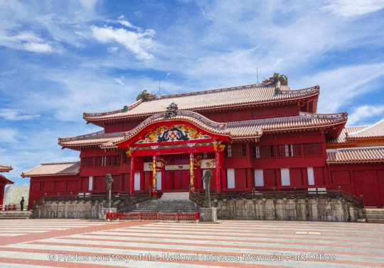 The majestic Seiden (Main Hall) of Shuri Castle, a vibrant red, Okinawan-style wooden building with a tiled plaza in front, set against a bright blue sky.