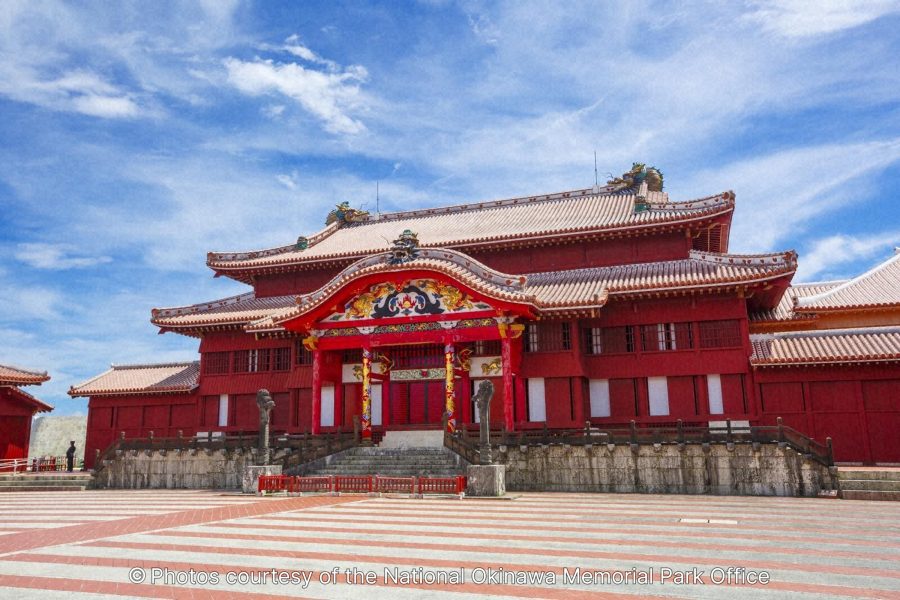 The majestic Seiden (Main Hall) of Shuri Castle, a vibrant red, Okinawan-style wooden building with a tiled plaza in front, set against a bright blue sky.