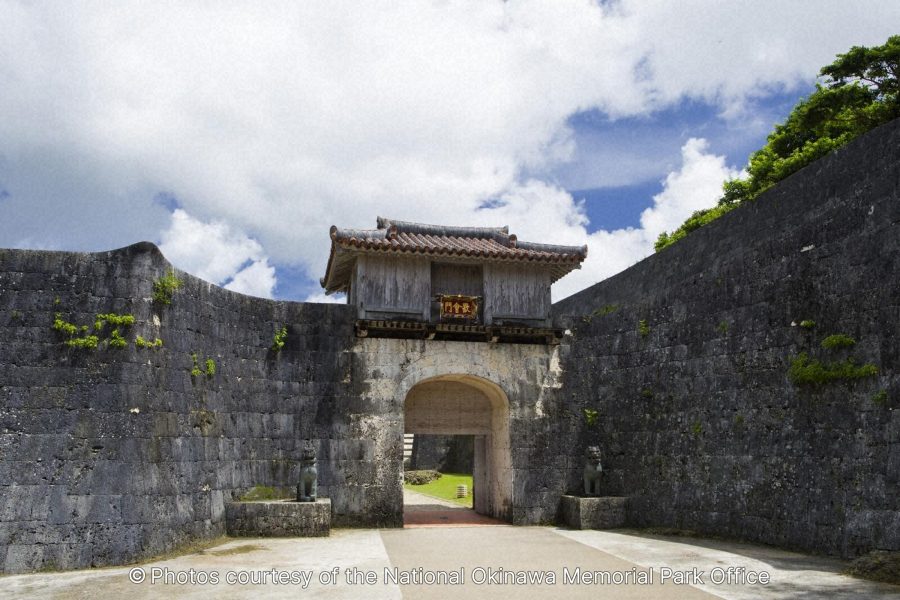 The Kankaimon Gate of Shuri Castle, an arched stone gate flanked by high stone walls with two stone Shisa statues on pedestals.