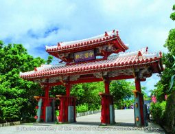 The Shureimon Gate, a bright red, traditional Ryukyu-style gate with a distinctive curved, red-tiled roof, surrounded by lush tropical greenery.