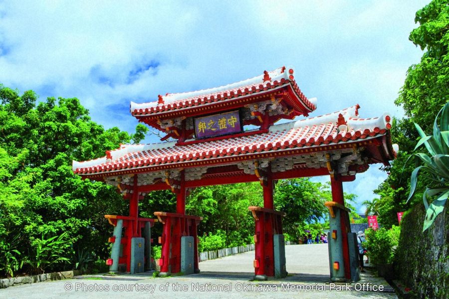 The Shureimon Gate, a bright red, traditional Ryukyu-style gate with a distinctive curved, red-tiled roof, surrounded by lush tropical greenery.