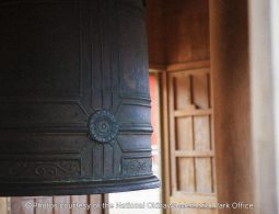 A close-up of a large, dark bronze temple bell with intricate circular patterns, hanging within a wooden structure.