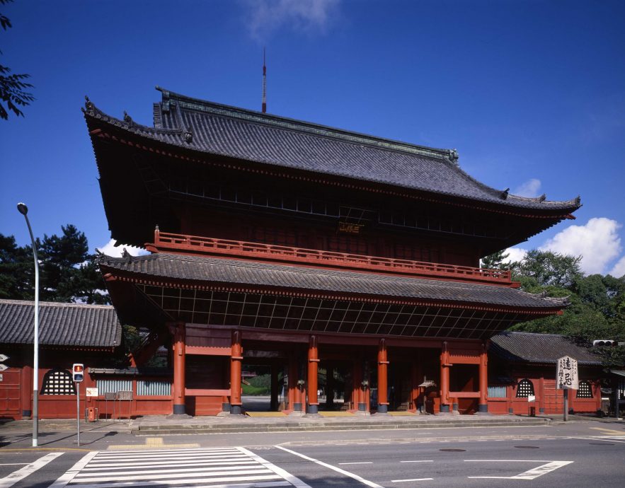 The towering red and black Sangedatsu-mon Gate (Main Gate) of Zojo-ji Temple, standing over a busy city street intersection.