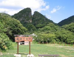 A large, distinctive split mountain peak known as Doyu-no-warito on Sado Island, viewed from a grassy field with a rustic wooden sign in the foreground.