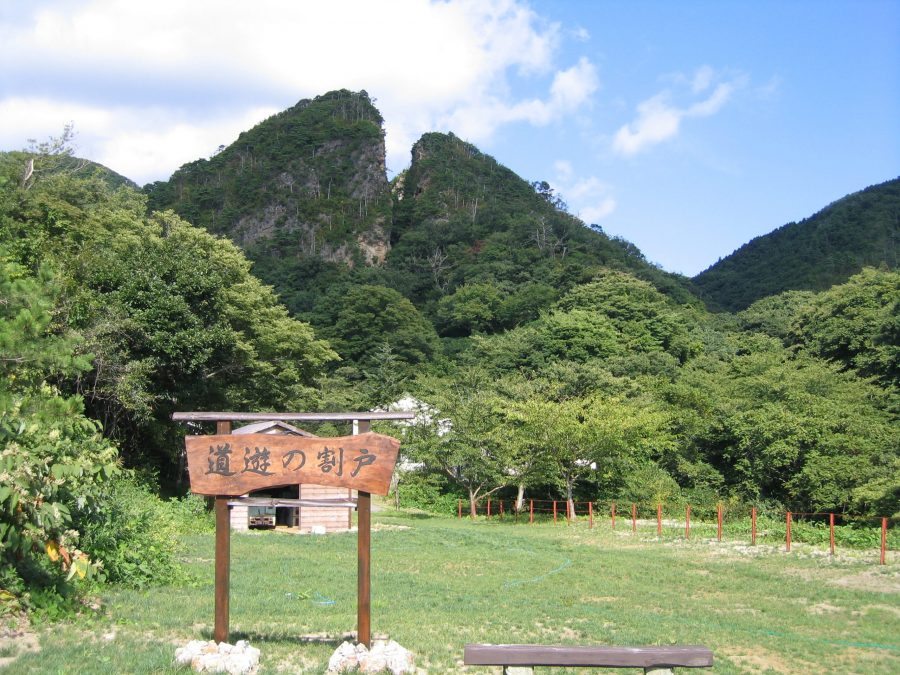 A large, distinctive split mountain peak known as Doyu-no-warito on Sado Island, viewed from a grassy field with a rustic wooden sign in the foreground.