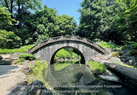 The Engetsukyō (Full Moon Bridge), a traditional stone arch bridge crossing a small, calm stream in a lush, tree-filled landscape.