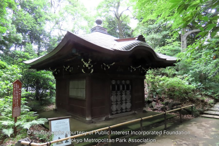 A small, traditional Japanese wooden shrine or structure with a curved roof, surrounded by dense green forest and mossy ground in the garden.