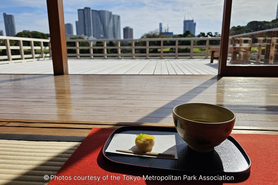 Close-up of matcha tea and a small wagashi sweet served on a black tray on a tatami mat floor inside a teahouse, with the modern city skyline visible through the open doorway.