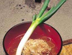 A bowl of Negi Soba (buckwheat noodles) from Ouchi-juku, served in a red lacquer bowl with bonito flakes and a single, large green leek used as a utensil, set against a rustic hearth background.