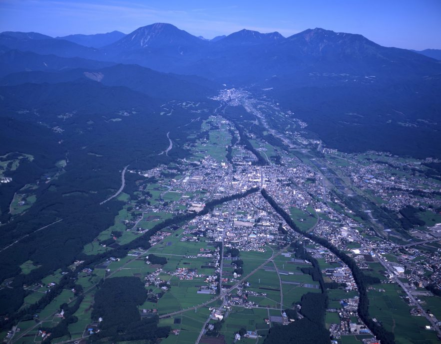 An aerial view of a city (likely Nikko or the surrounding area) nestled in a valley, surrounded by green mountains and fields.