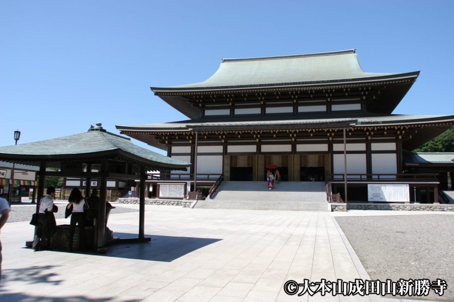 The modern Great Main Hall (Dai Hondo) of Naritasan Shinsho-ji Temple under a bright blue sky.
