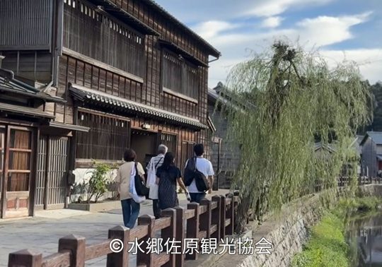 Traditional wooden merchant houses line the banks of a canal in Sawara, Chiba, with a willow tree hanging over the water and people walking along the historic street.
