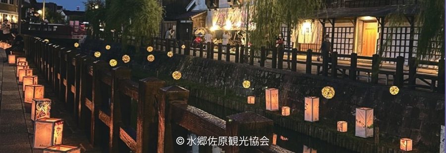 A canal in Sawara illuminated at night by glowing paper lanterns placed along the stone wall and wooden fence, reflecting light on the water.