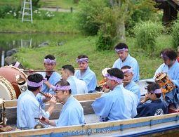 A group of men in light blue festival attire and headbands playing traditional drums and flutes on a boat on the water in Sawara.