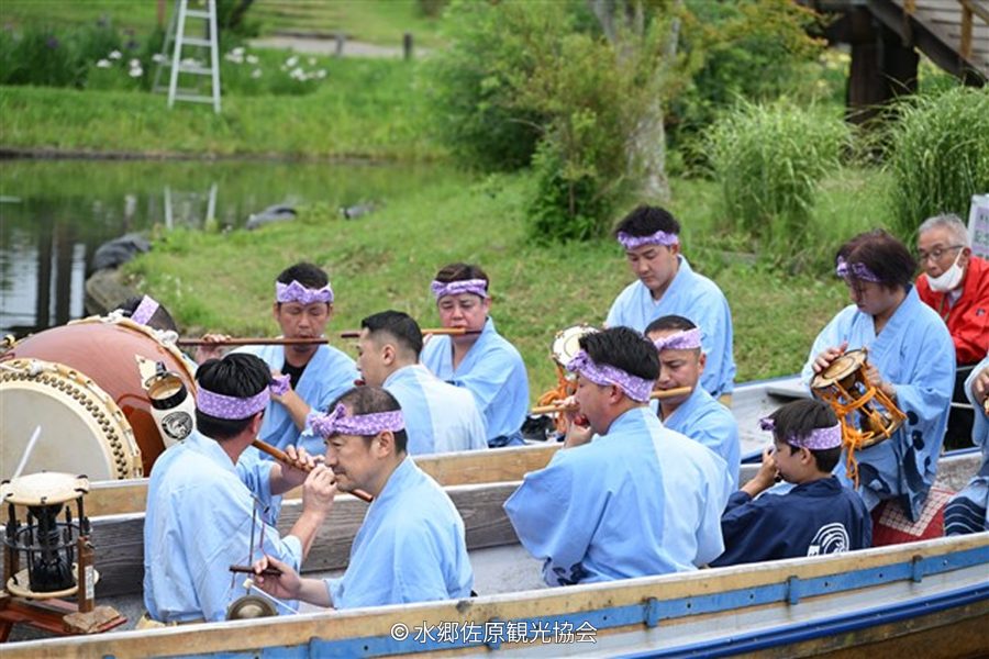 A group of men in light blue festival attire and headbands playing traditional drums and flutes on a boat on the water in Sawara.