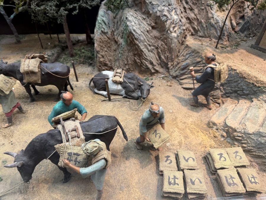 Diorama showing Edo-period workers at Sado Gold Mine loading sacks onto cows for transport, with more sacks labeled with Japanese kana lying on the ground.