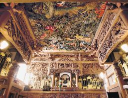 Intricate, colorful ceiling carving by Ishikawa Uncho in a temple interior, featuring dragons and elaborate cloud motifs above a richly carved altar.