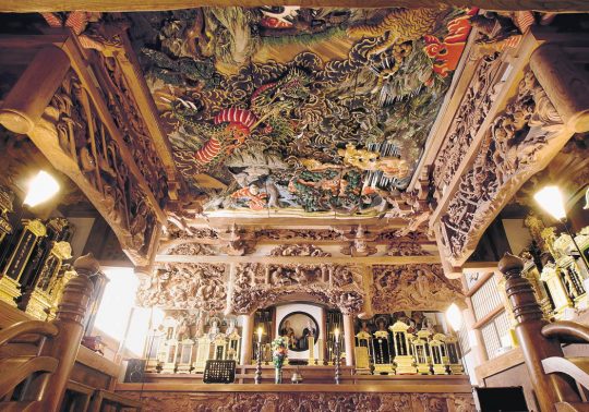 Intricate, colorful ceiling carving by Ishikawa Uncho in a temple interior, featuring dragons and elaborate cloud motifs above a richly carved altar.