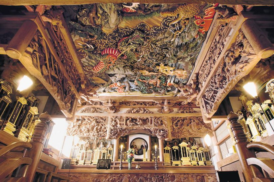 Intricate, colorful ceiling carving by Ishikawa Uncho in a temple interior, featuring dragons and elaborate cloud motifs above a richly carved altar.