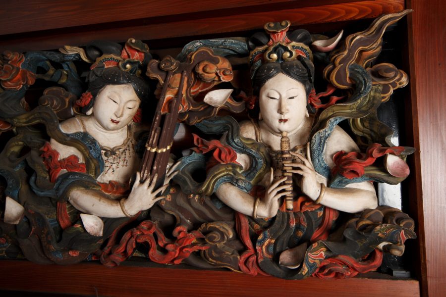 A close-up of a richly painted and carved wooden panel depicting two female figures in elaborate robes, possibly celestial maidens (tennyo).