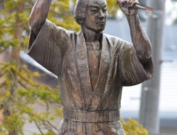 A bronze statue of a man (presumably Uncho) holding a mallet and chisel, posed mid-carving, set outdoors against green and yellow foliage.