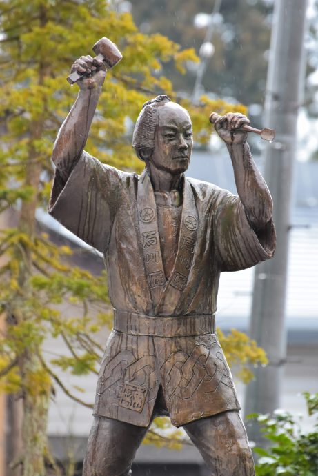 A bronze statue of a man (presumably Uncho) holding a mallet and chisel, posed mid-carving, set outdoors against green and yellow foliage.