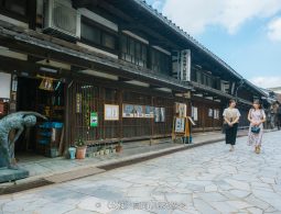 Traditional Japanese wooden merchant houses lining a stone-paved street, with two women walking past a copper statue.