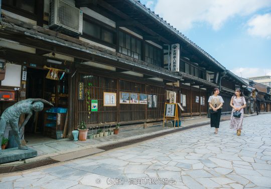 Traditional Japanese wooden merchant houses lining a stone-paved street, with two women walking past a copper statue.