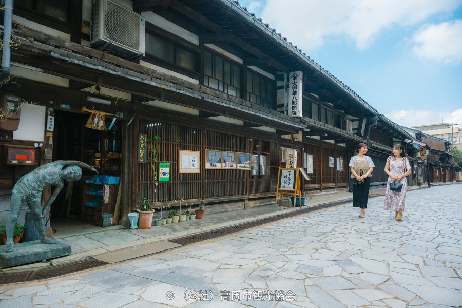 Traditional Japanese wooden merchant houses lining a stone-paved street, with two women walking past a copper statue.
