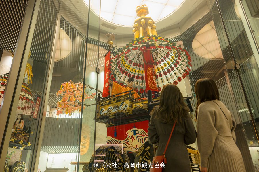 Museum visitors looking up at a richly decorated, towering festival float (Mikuruma-yama) displayed behind glass.