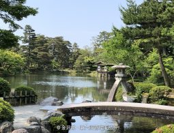 The S-shaped bridge and stone lantern (likely Kotoji Lantern) on Kasumigaike Pond in Kenroku-en Garden, surrounded by lush summer greenery.