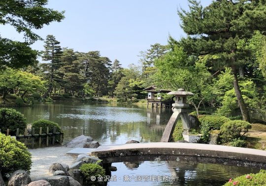 The S-shaped bridge and stone lantern (likely Kotoji Lantern) on Kasumigaike Pond in Kenroku-en Garden, surrounded by lush summer greenery.