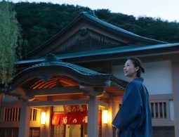A woman wearing a dark kimono jacket and patterned skirt stands smiling in front of a traditional Japanese public bathhouse entrance at dusk in Yamanaka Onsen.