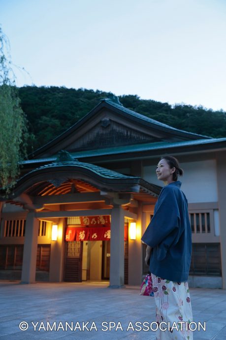 A woman wearing a dark kimono jacket and patterned skirt stands smiling in front of a traditional Japanese public bathhouse entrance at dusk in Yamanaka Onsen.