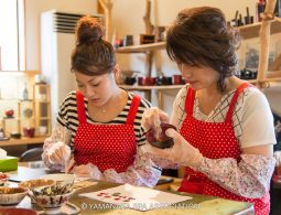 Two women in matching red polka-dot aprons are hand-painting and decorating small bowls, likely part of a traditional Yamanaka lacquerware craft workshop.
