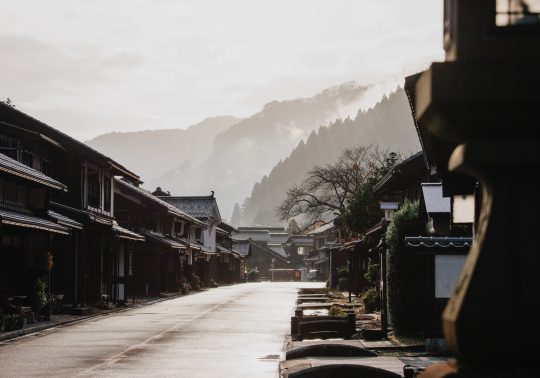 A view down the street of Kumagawa-juku in the early morning, with traditional houses on both sides and foggy, tree-covered mountains in the distance.