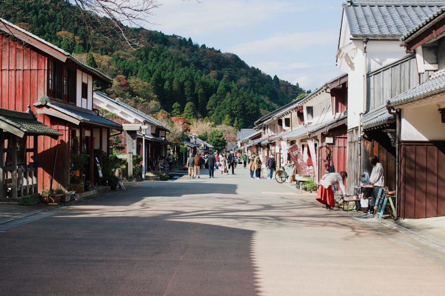 The main street of Kumagawa-juku filled with people, showcasing houses with contrasting white and reddish-brown facades against a backdrop of dense forest.
