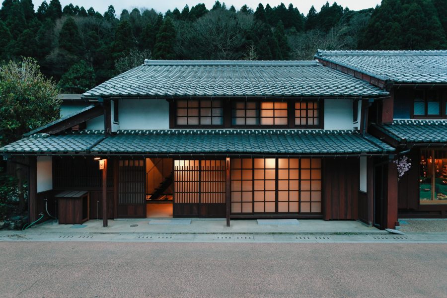 A traditional two-story Japanese house in Kumagawa-juku at dusk, with light glowing warmly from the paper and wood-paneled windows.